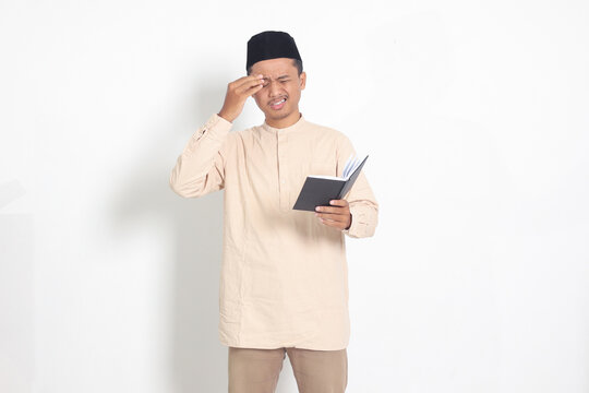 Portrait of confused muslim man in koko shirt with peci reading a notebook. Unhappy Asian guy holding his forehead, having headache. Isolated image on white background