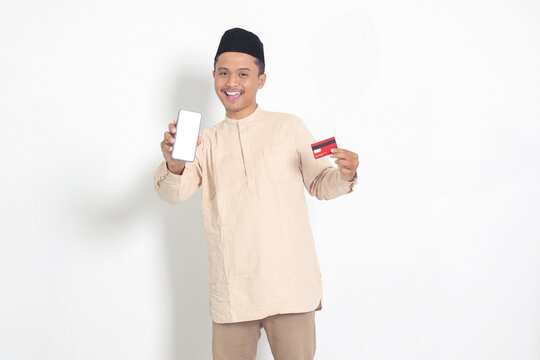 Portrait of attractive Asian muslim man in koko shirt with skullcap holding a credit card while showing empty screen on mobile phone for mock up and graphic. Isolated image on white background