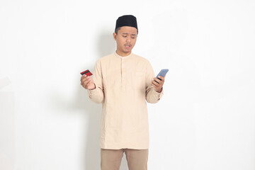 Portrait of upset Asian muslim man in koko shirt with skullcap holding a mobile phone and...