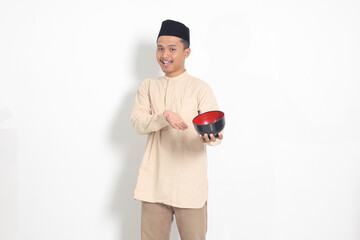 Portrait of excited Asian muslim man in koko shirt with skullcap showing and holding an empty...