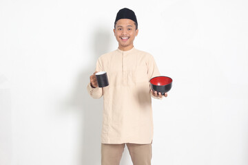 Portrait of excited Asian muslim man in koko shirt with skullcap holding a mug and empty bowl. Bowl...