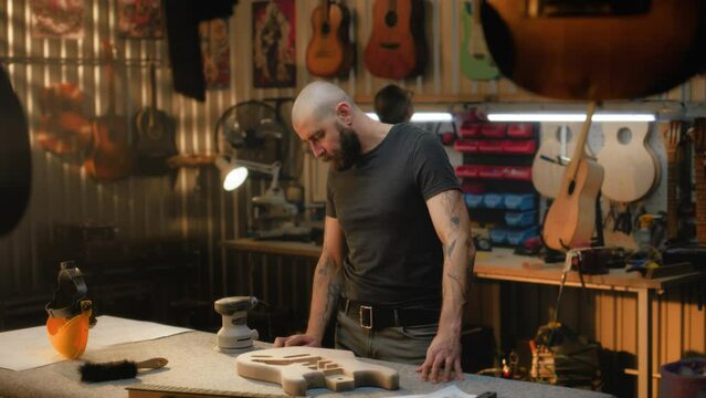 Middle aged tattooed craftsman stands near workplace. Caucasian man looks at guitars and starts working. Professional carpenter works in light stylish workshop. Handmade and entrepreneurship concept.