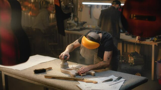 Middle aged tattooed carpenter in protective mask grinds guitar body using grinding machine. Caucasian artisan makes electro guitar in light stylish workshop. Handcraft and small business concept.