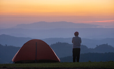 Fototapeta na wymiar Traveller is standing by the tent during overnight camping while looking at the beautiful scenic sunset over the mountain for outdoor adventure vacation travel concept