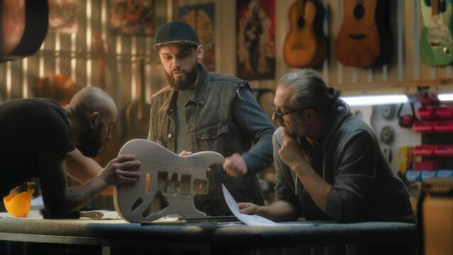Mature Caucasian carpenter looks at blueprints and discusses work in stylish light workshop. Male tattooed craftsman holds guitar body and talks with colleagues. Handmade and entrepreneurship concept.