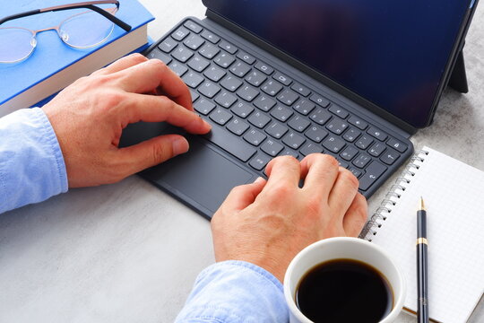 コーヒーを飲みながら、キーボードを接続したタブレットPCでデスクワークをする男性の両手のイメージ画像
