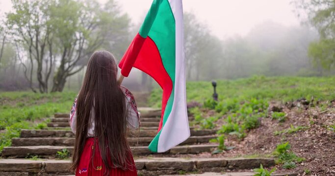 Bulgarian girl standard bearer in ethnic folklore costume with bulgarian flag climbs the stairs toward peak Shipka monument, 3th March Liberation day of Bulgaria
