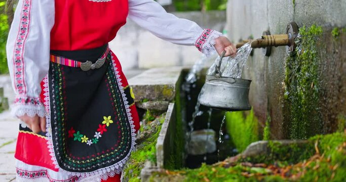 Bulgarian woman in ethnic folklore embroidery costume filling water from traditional forest stone drinking mineral water fountain video