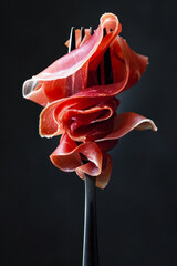  Elegantly twirled slice of prosciutto ham on an fork, isolated on a dark background