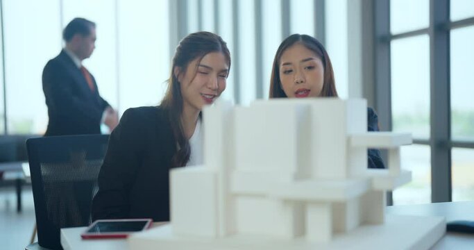 Young Asian woman presents a building model to team at the office. They discuss building design together in the office.