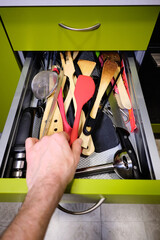 Unrecognizable man's hand taking a wooden spoon inside a kitchen drawer full of knives and kitchen utensils.