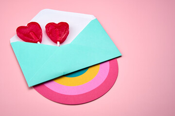 Representation of a love letter with two red heart-shaped lollipops inside and a rainbow, on a pink background