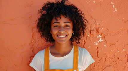 Young woman with curly hair smiling wearing a white t-shirt and orange suspenders standing against...