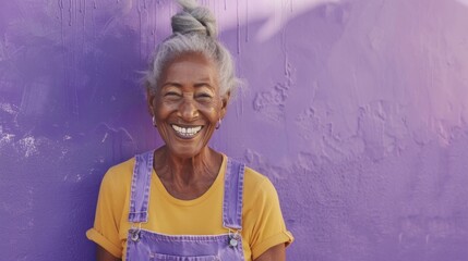 A joyful elderly woman with gray hair wearing a yellow shirt and purple overalls smiling against a vibrant purple wall.