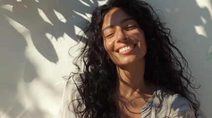 Beautiful woman with long curly hair smiling and enjoying the sun with her eyes closed against a white wall with shadows of leaves.