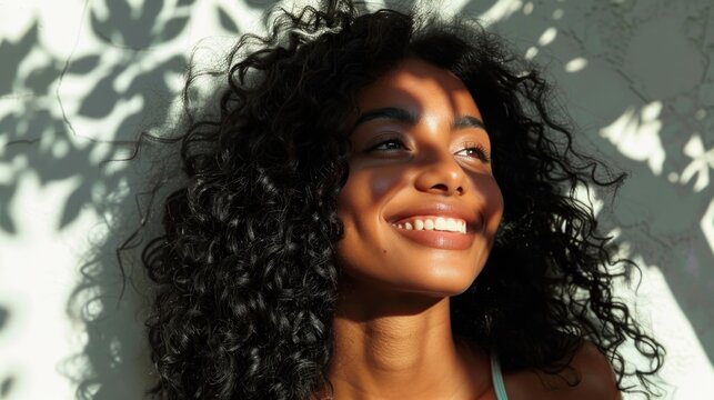 A joyful woman with curly hair smiling at the camera with her face partially shaded by sunlight and leaf shadows.