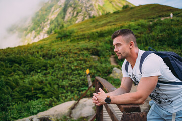 A man in light-colored clothes leans on a wooden fence. Against the background of mountains covered...