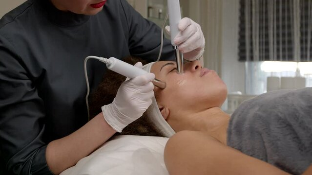 Close-up of professional cosmetologist making anti aging procedure and microcurrent facial therapy to lovely African American woman, lifting and tightening face muscles at beauty salon,