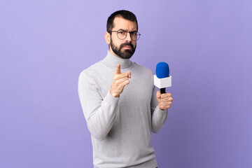 Adult reporter man with beard holding a microphone over isolated purple background frustrated and...