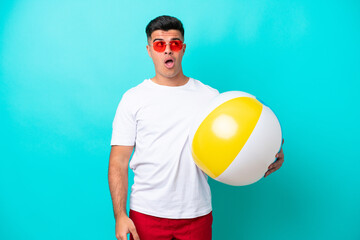 Young caucasian man holding a beach ball isolated on blue background looking up and with surprised...