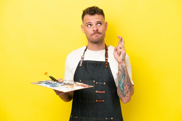 Young artist caucasian man holding a palette isolated on yellow background with fingers crossing...