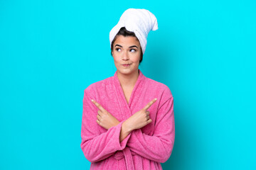 Young Brazilian woman with a bathrobe isolated on blue background pointing to the laterals having...