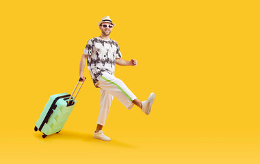 Happy male tourist goes on summer vacation and travels abroad. Cheerful young man in T shirt, white trousers, hat and sunglasses with green holiday suitcase walking isolated on yellow color background