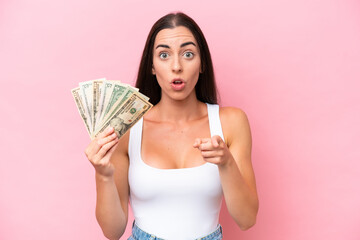 Young caucasian woman taking a lot of money isolated on pink background surprised and pointing front