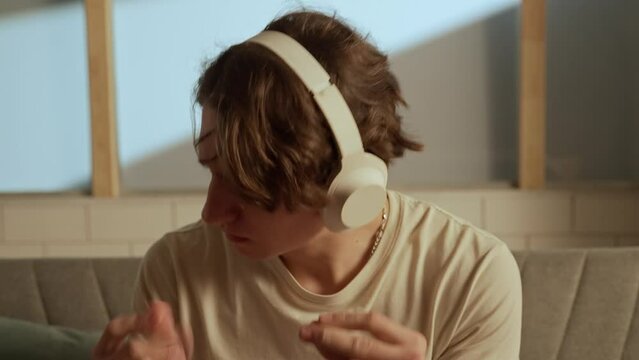 Portrait of adult person sitting on the couch. Close up young man in headphones listening to music, singing song jamming to the song clapping.