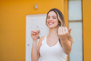 Young pretty woman holding home keys at outdoors inviting to come with hand. Happy that you came