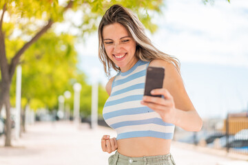 Young pretty woman at outdoors using mobile phone and doing victory gesture