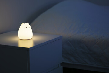 Cute cat shaped night lamp standing on a bedside table next to bed. Bedside lamp. Night lamp standing next to bed. Bedroom lamp on a night table next to a sleeping bed in a dark room. 