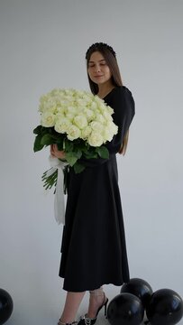 Sophisticated Lady with White Roses. Woman in elegant black dress holds large bouquet of white roses, surrounded by black balloons on floor.