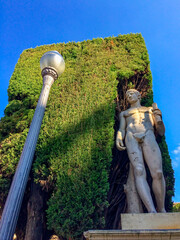 The image shows a statue and a street lamp near a wall covered in greenery under a clear sky.