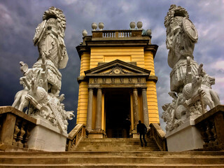 The image shows a person ascending steps towards a neoclassical building flanked by two intricate statues under a stormy sky.