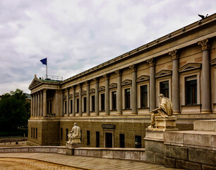 Fototapeta na wymiar A classical building with columns, statues, and a flag under a cloudy sky. Austrian parliament building, Austrian Parliament, cloudy weather, sculptures, work of architectural art, authorities
