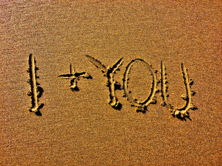 The image displays ‘I + YOU’ deeply etched into a sandy surface.