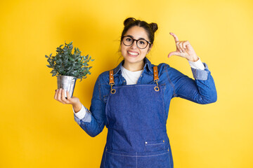 Young caucasian gardener woman holding a plant isolated on yellow background smiling and confident...