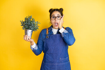 Young caucasian gardener woman holding a plant isolated on yellow background afraid and shocked,...
