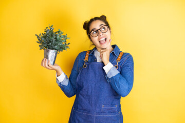 Young caucasian gardener woman holding a plant isolated on yellow background happy face thinking...