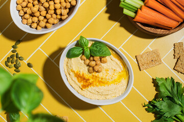 Delicious chickpea hummus in a plate on a bright yellow background with fresh vegetables. 