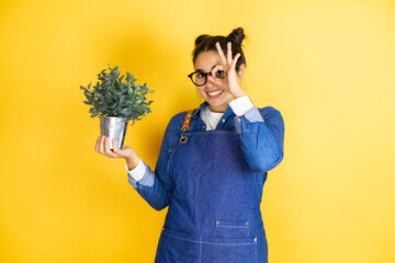 Young caucasian gardener woman holding a plant isolated on yellow background doing ok gesture...