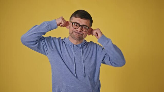 Playful senior man in glasses making monkey faces on yellow background