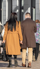 Fototapeta premium Back view of fashionable couple wearing brown coats walking on the street holding hands in sunny day. Rear view of stylish young couple in love walking on the street in the city center. City life