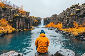 Person in yellow jacket seated by autumnal river, basalt columns and waterfall in distance. Solitary figure contemplates at riverbank, autumn hues frame the peaceful waterfall scene