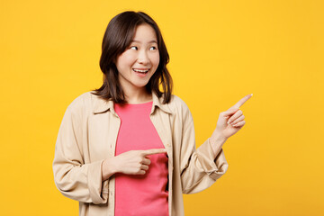 Young happy woman of Asian ethnicity she wear pink t-shirt beige shirt pastel casual clothes point index finger aside on area isolated on plain yellow background studio portrait. Lifestyle portrait.