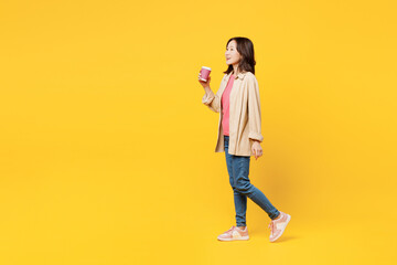 Full body young woman of Asian ethnicity wear pink t-shirt beige shirt casual clothes hold takeaway delivery craft paper brown cup coffee to go isolated on plain yellow background. Lifestyle portrait.