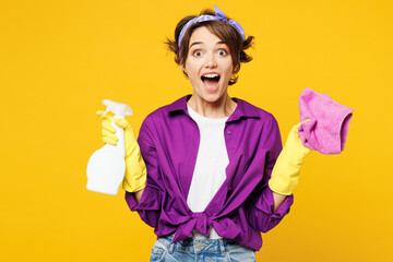 Young surprised shocked woman wears purple shirt rubber gloves do housework tidy up hold in hand rag spray bottle look camera isolated on plain yellow background studio portrait. Housekeeping concept. - obrazy, fototapety, plakaty