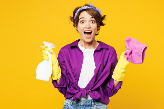 Young surprised shocked woman wears purple shirt rubber gloves do housework tidy up hold in hand rag spray bottle look camera isolated on plain yellow background studio portrait. Housekeeping concept.