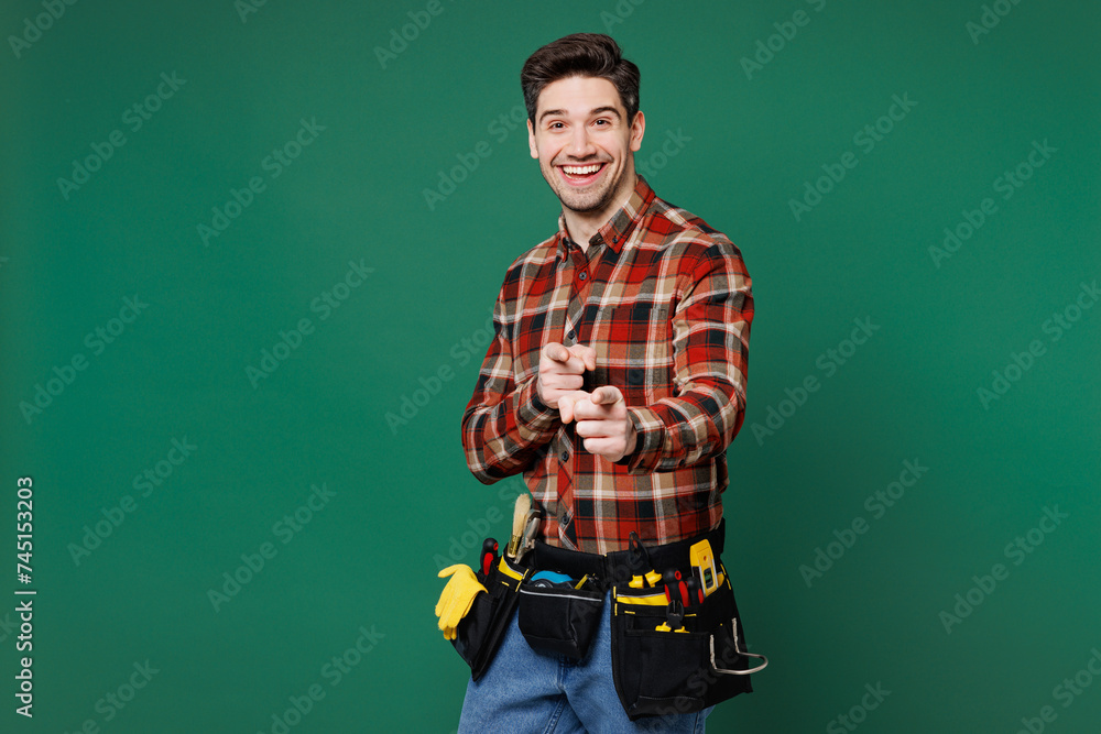 Wall mural Young happy employee laborer handyman man wear red shirt point index finger camera on you isolated on plain green background. Instruments accessories for renovation apartment room Repair home concept